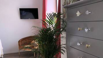Huoneessa on harmaa lipasto koristeellisilla nupeilla, ruukussa oleva kasvi, puinen tuoli ja seinälle asennettu televisio.