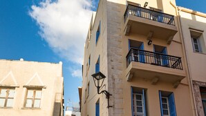 Une rue étroite, bordée de bâtiments de chaque côté, avec des balcons, des plantes en pot et un ciel d’un bleu éclatant.