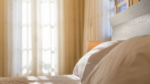 Un lit bien fait, avec une couette blanche et des oreillers, une tête de lit en bois et des rideaux légers qui laissent passer la lumière naturelle.