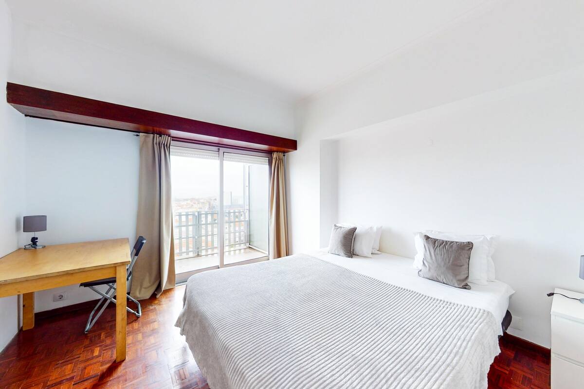 Une chambre d’hôtel avec un lit, un bureau, une chaise, une lampe et une fenêtre avec des rideaux.