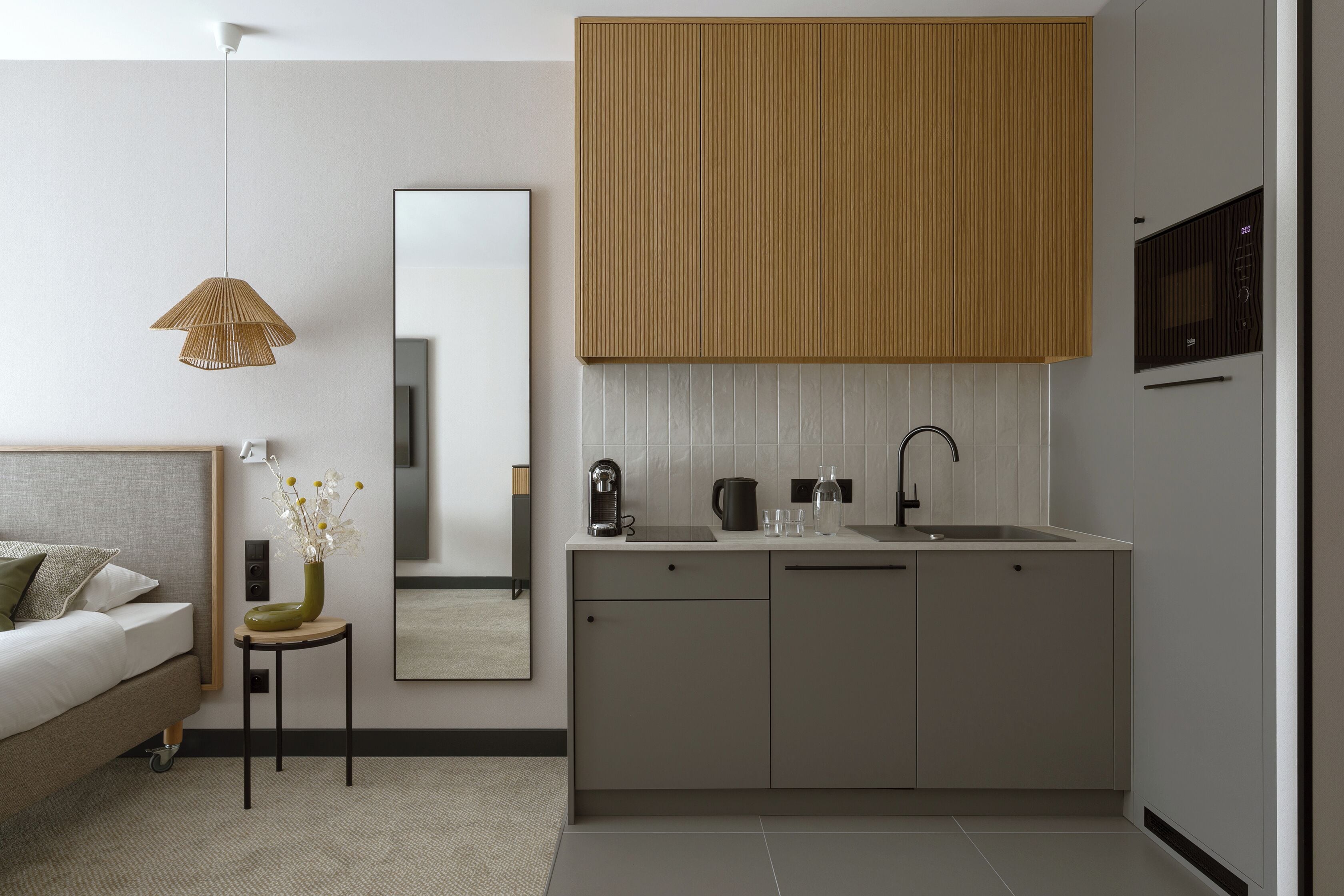 A modern bedroom with a bed, bedside table, and a small table with a vase. Adjacent is a minimalist kitchen with a sink, cabinets, and a mirror.