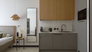 A modern bedroom with a bed, bedside table, and a small table with a vase. Adjacent is a minimalist kitchen with a sink, cabinets, and a mirror.