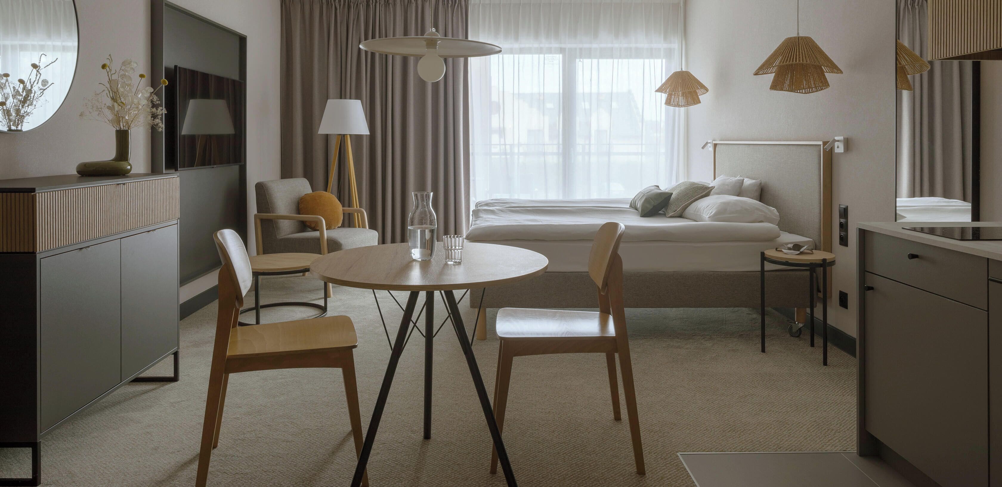 Una habitación de hotel moderna con cama, mesita de noche, lámpara y un rincón de comedor con mesa y sillas.