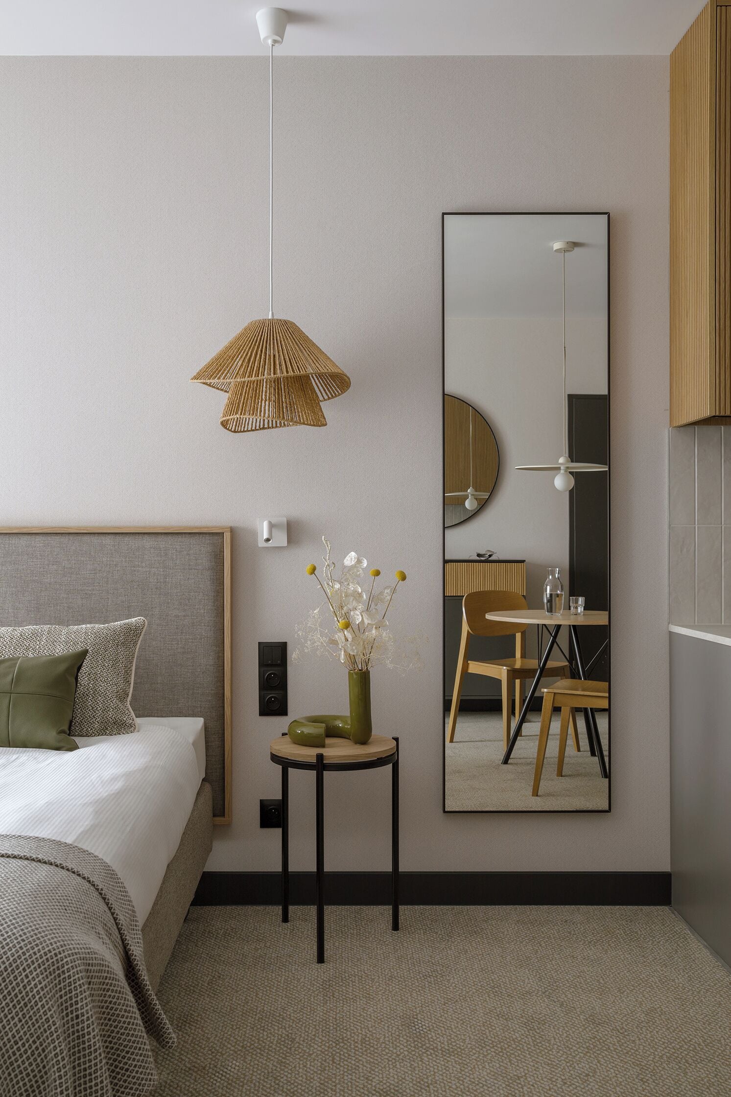 A modern hotel room with a large bed, a small round table with a vase of flowers, a large mirror, and a hanging light fixture.