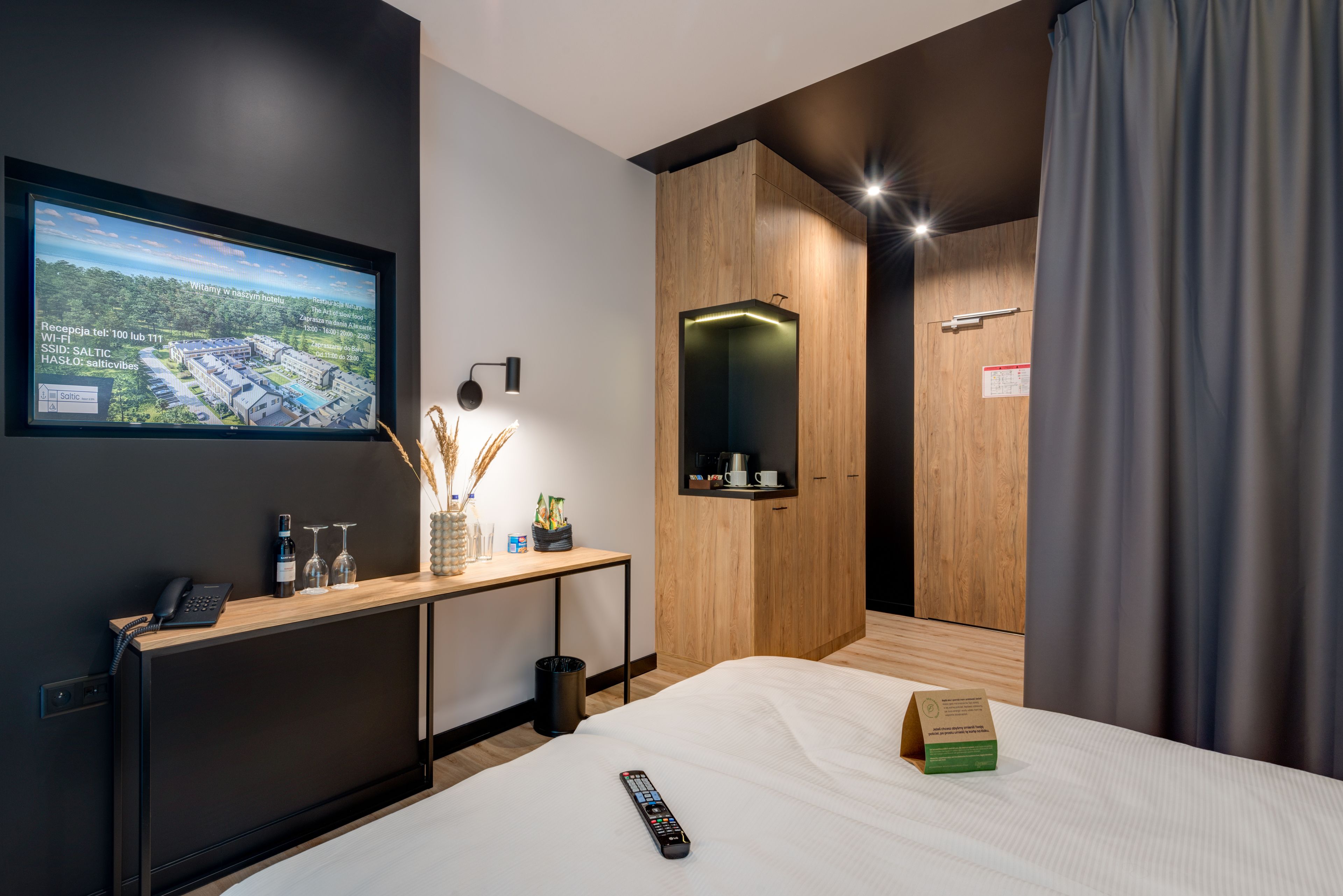 Une chambre d’hôtel moderne équipée d’un lit, d’un bureau avec un téléphone et des verres à vin, d’un téléviseur diffusant un paysage pittoresque et d’une armoire en bois.