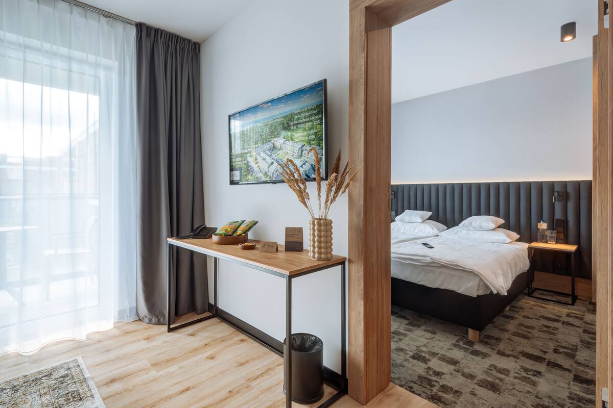 A modern hotel room with a wooden desk, a bed with a grey headboard, a framed picture on the wall, and a window with grey curtains.