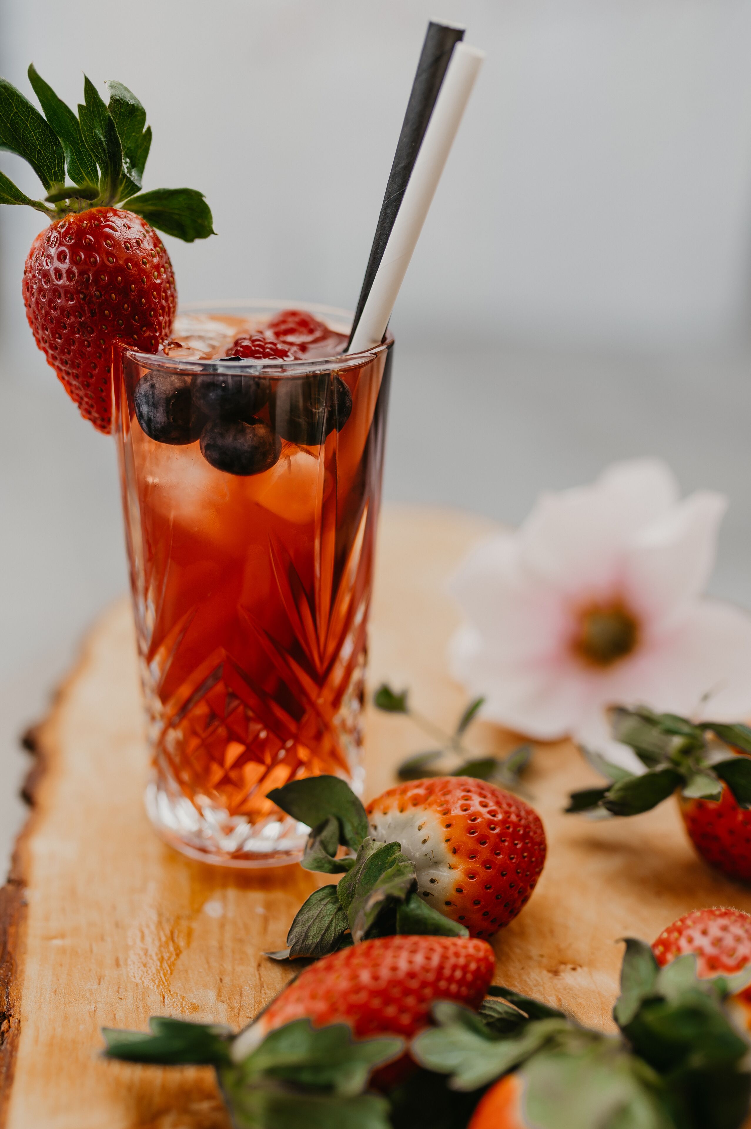 A glass of iced beverage with strawberries and blueberries, placed on a wooden surface with more strawberries around.