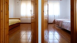 Una habitación con suelo de madera, una cama con colchón blanco y ropa de cama marrón, una ventana con cortinas translúcidas y una puerta de madera.