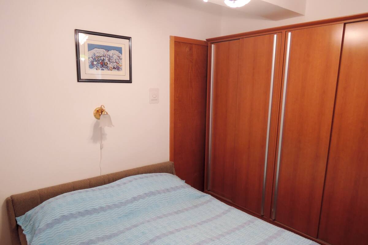 A bedroom with a bed, a wooden wardrobe, a framed picture on the wall, and a wall-mounted lamp.
