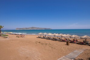 A beach with rows of sun loungers and thatched umbrellas, a wooden walkway, and a view of the sea and a distant island. - Steps to the Beach Spacious Home-Parking (Chania)