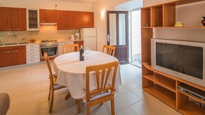 A modern kitchen with wooden cabinets, a dining table with chairs, a built-in oven, and a large flat-screen TV on a wooden stand. - Apartment Anica (Malinska-Dubasnica)
