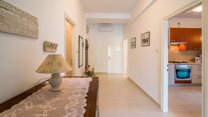 A hallway with a wooden table, a lace table runner, and a lamp. There are framed pictures on the walls and a door leading to a kitchen area with wooden cabinets and a stove. - Apartment Anica (Malinska-Dubasnica)