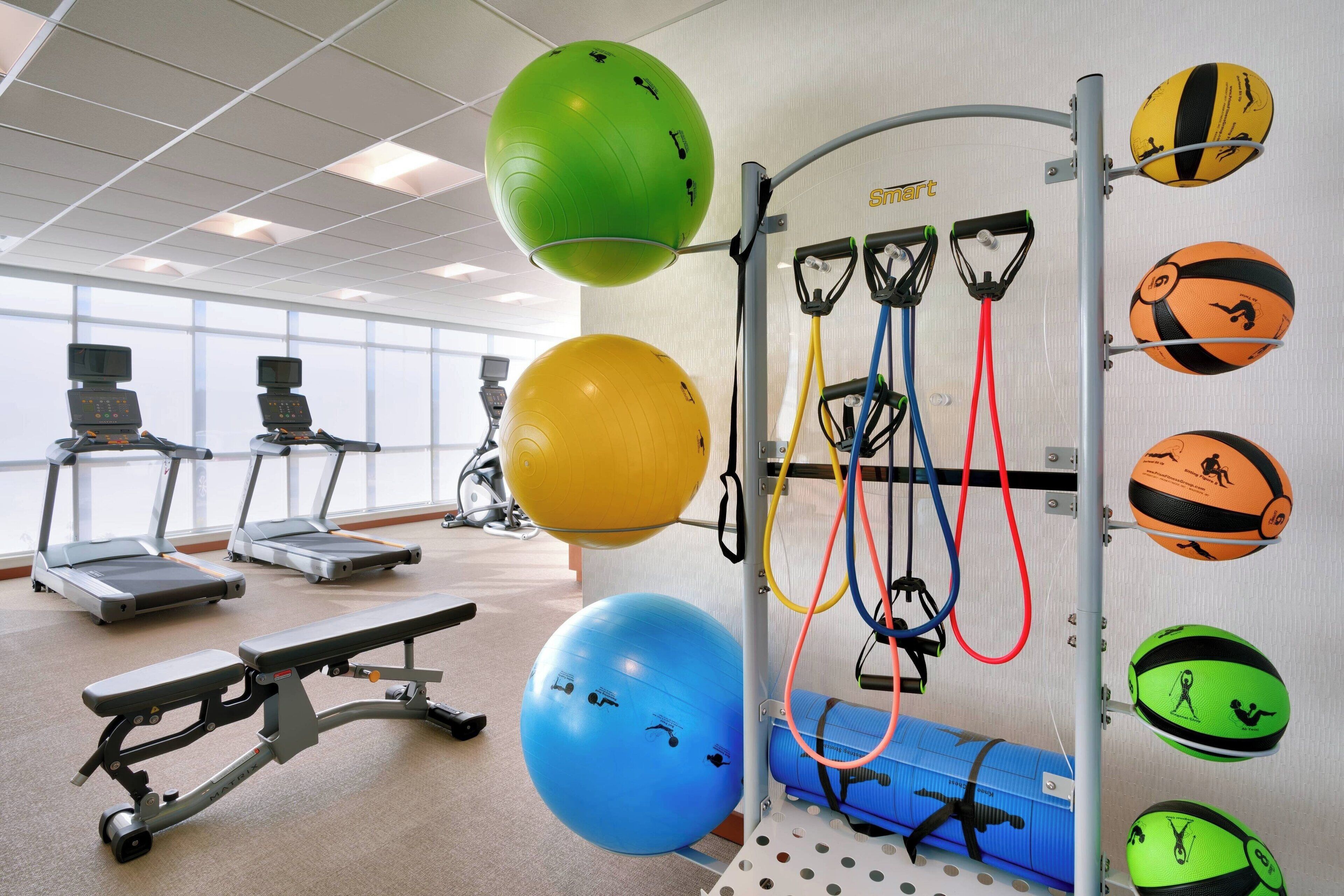 Un gimnasio con equipo de ejercicio, cintas de correr y un estante de pared con pelotas de ejercicio y bandas de resistencia.