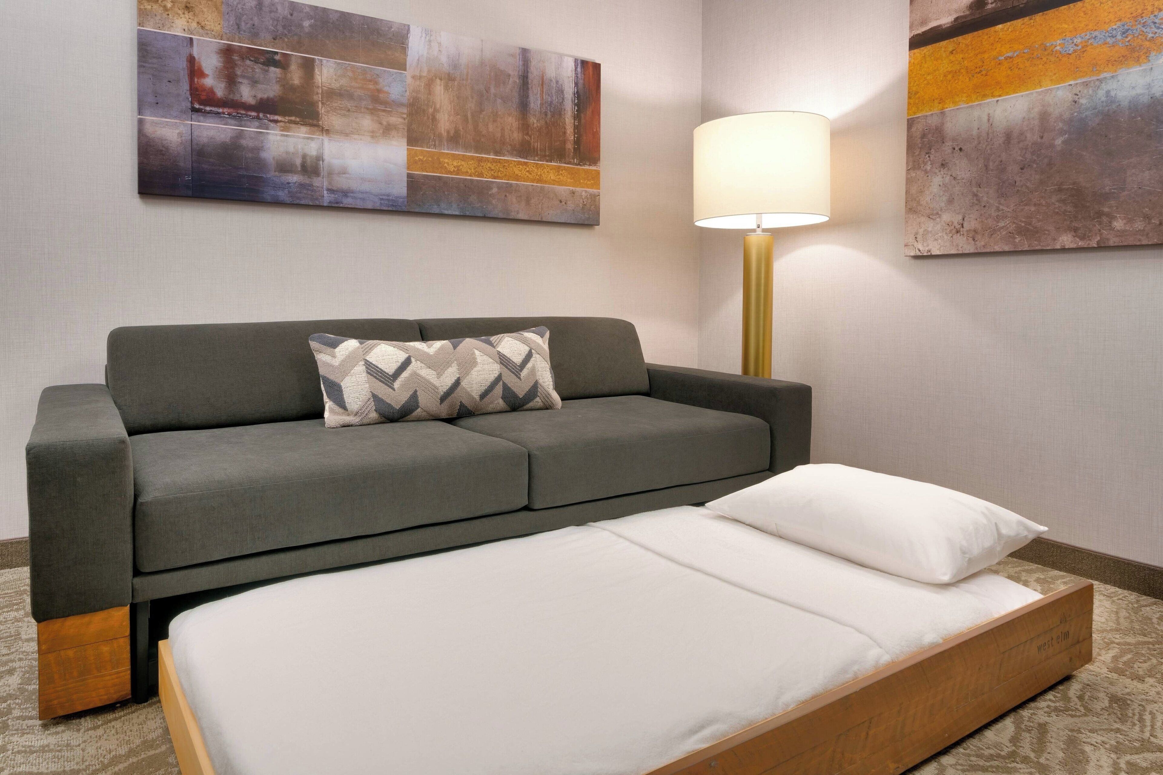 Quarto de hotel moderno com sofá cinzento, estrutura de cama de madeira e uma grande pintura abstrata na parede.