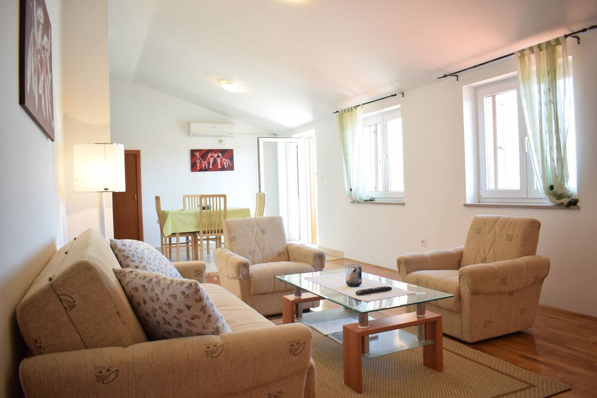 Ein Wohnzimmer mit beigefarbenen Sofas, einem Glastisch und einem Essbereich im Hintergrund.