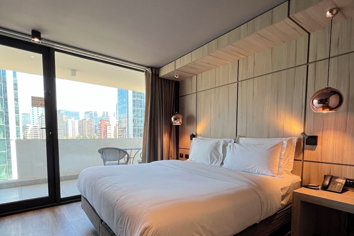 A modern hotel room with a large bed, a desk with a phone, and a view of the city through large windows.