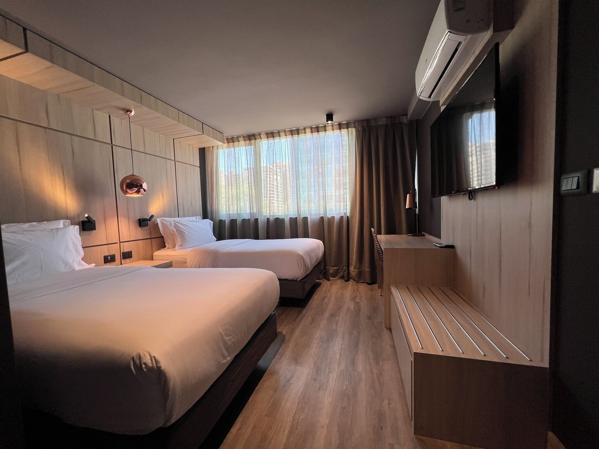 A hotel room with two beds, a desk, a TV, and an air conditioning unit.