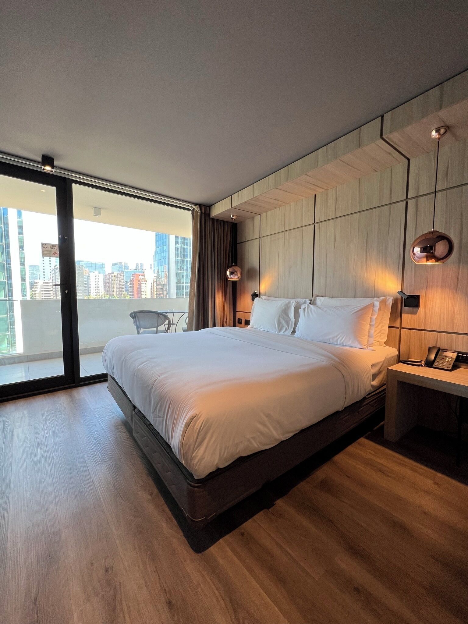 A modern hotel room with a large bed, a desk with a phone, and a view of the city through large windows.