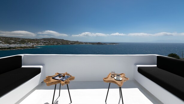 Un balcón con dos mesas de madera, cojines negros y vista al mar y la costa distante.