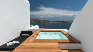 Un balcón privado con un jacuzzi de madera, una silla de lounge y un libro, con vistas a un pueblo costero y al mar.