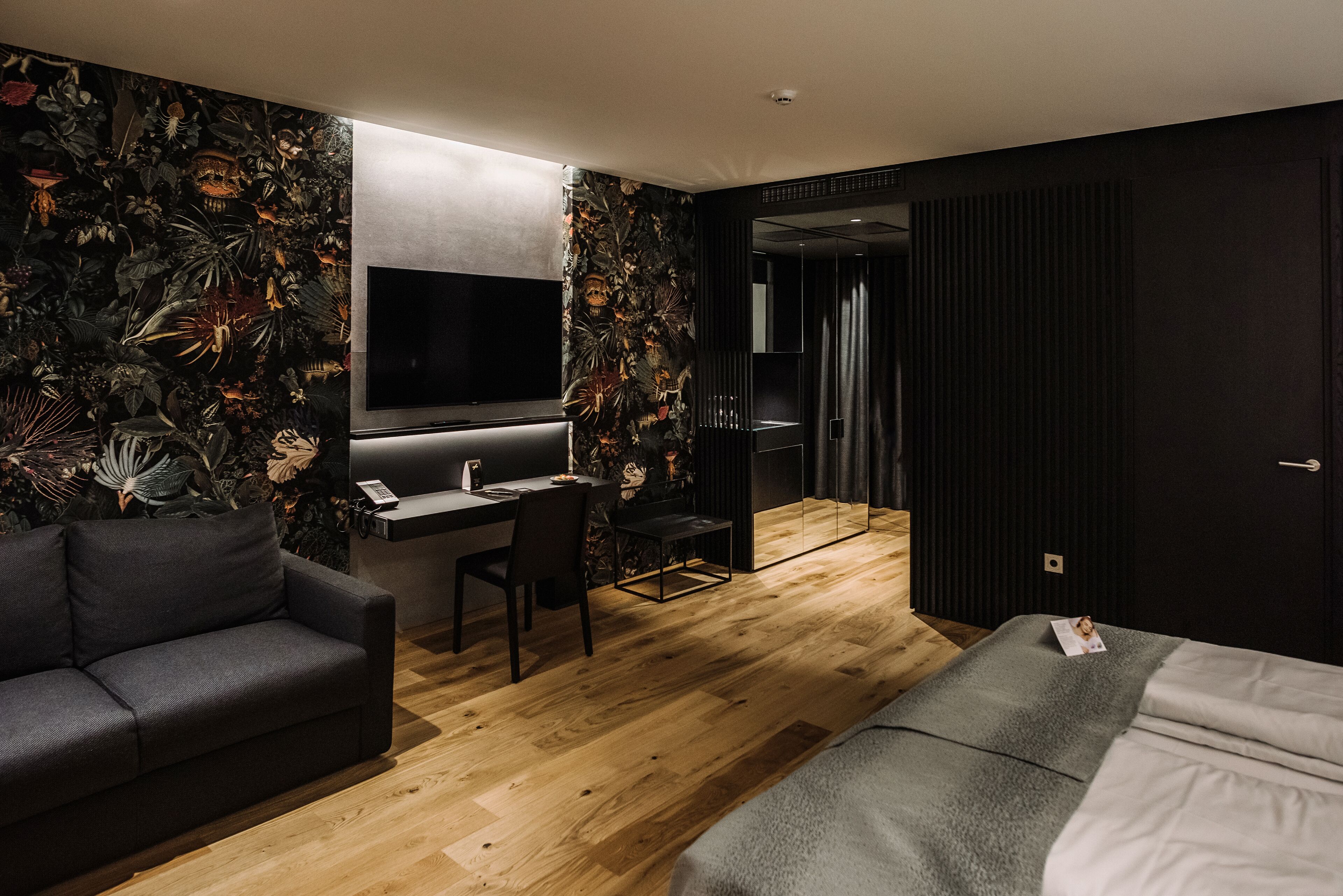 Une chambre d’hôtel moderne avec un plancher en bois, un canapé, un lit, un bureau avec une chaise, un téléviseur à écran plat et un grand miroir.