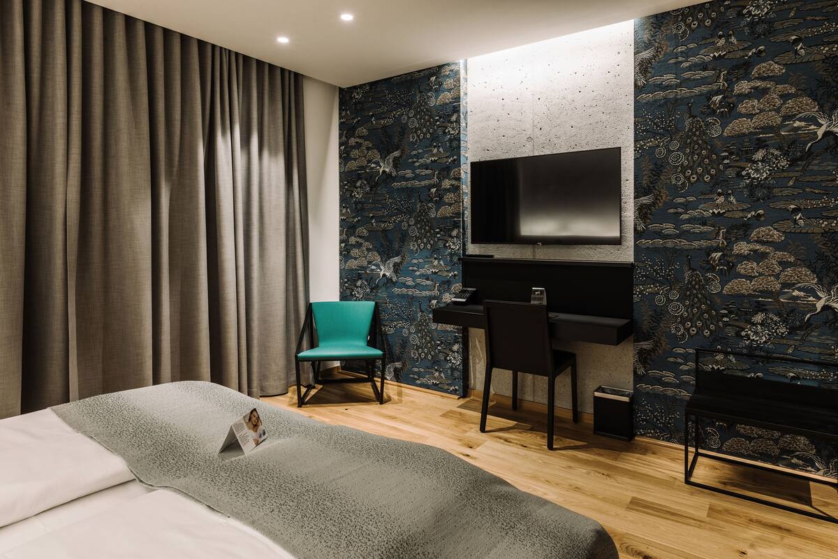 Ein modernes Hotelzimmer mit einem großen Bett, einem Schreibtisch mit Stuhl, einem flachbildfernseher an der Wand und einer Wandtapete mit Muster.