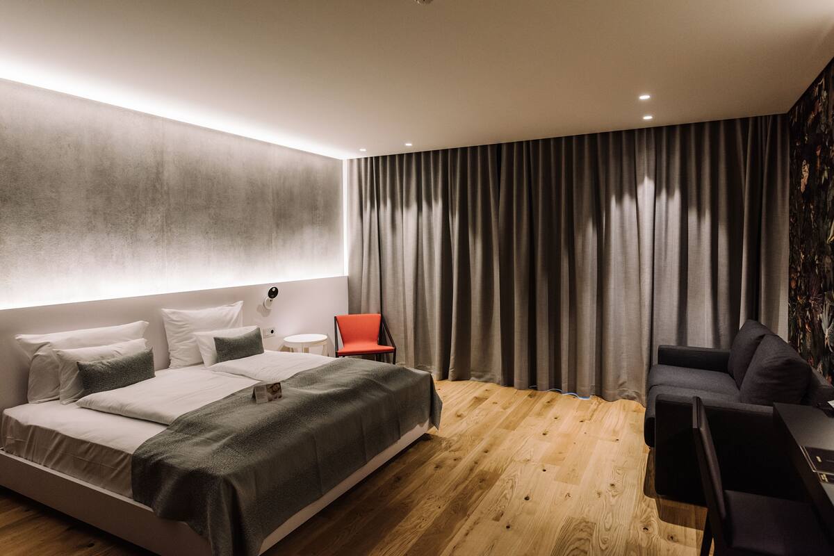 Ein modernes Schlafzimmer mit einem Bett, einem roten Sessel, einer Couch und einem großen Fenster mit Vorhängen.