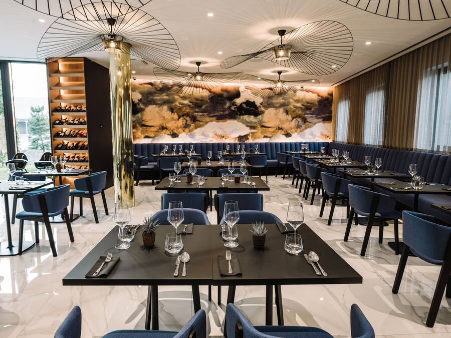 A modern restaurant with marble flooring, blue upholstered booths, and a wine cellar visible through a glass wall.