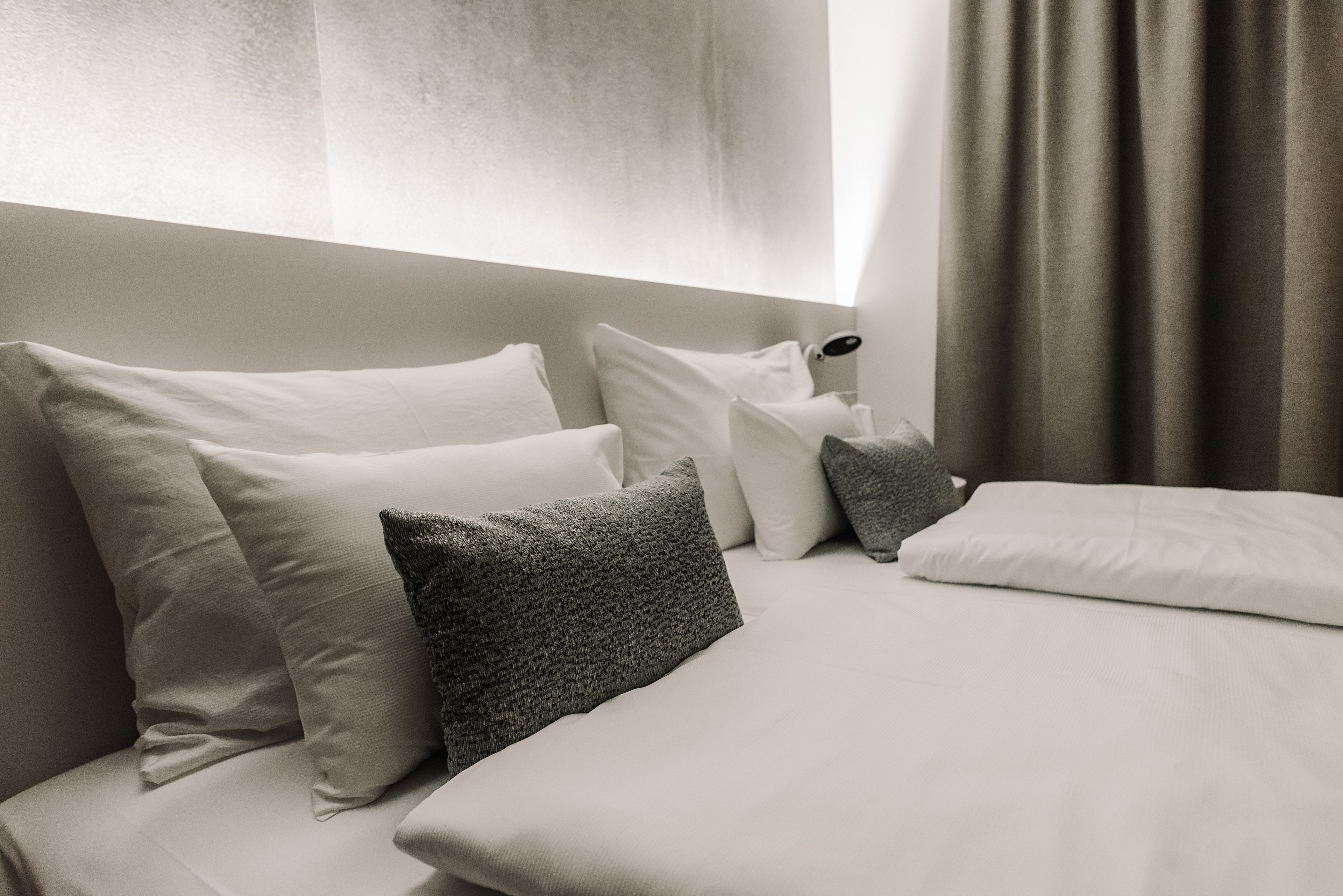 A neatly made bed with white and gray pillows, a bedside lamp, and a window with curtains.