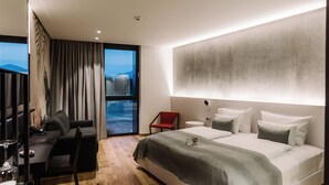 Una habitación de hotel moderna con una cama grande, un escritorio con una silla, una mesita redonda y vista al mar a través de un ventanal amplio.