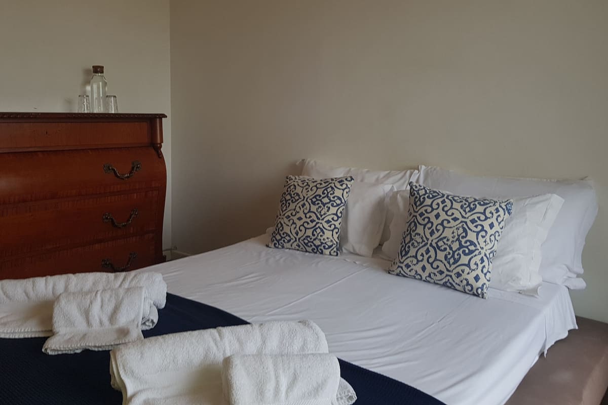 A neatly made bed with white linens and patterned pillows, a wooden dresser with drawers, and a blue throw blanket on the bed.