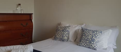 A neatly made bed with white linens and patterned pillows, a wooden dresser with drawers, and a blue throw blanket on the bed.
