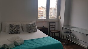 Un lit bien fait, recouvert d’une couverture turquoise, agrémenté de deux coussins à motifs et de serviettes pliées. Une chaise en bois et une petite table avec une lampe se trouvent à côté du lit. Une fenêtre donne sur un paysage urbain.