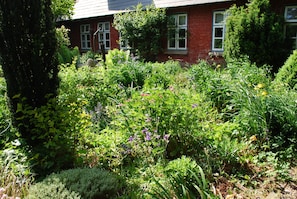 A garden with various plants and flowers in front of a brick building with windows. - Hotel - Pension ALTES PASTORAT (Langenhorn)