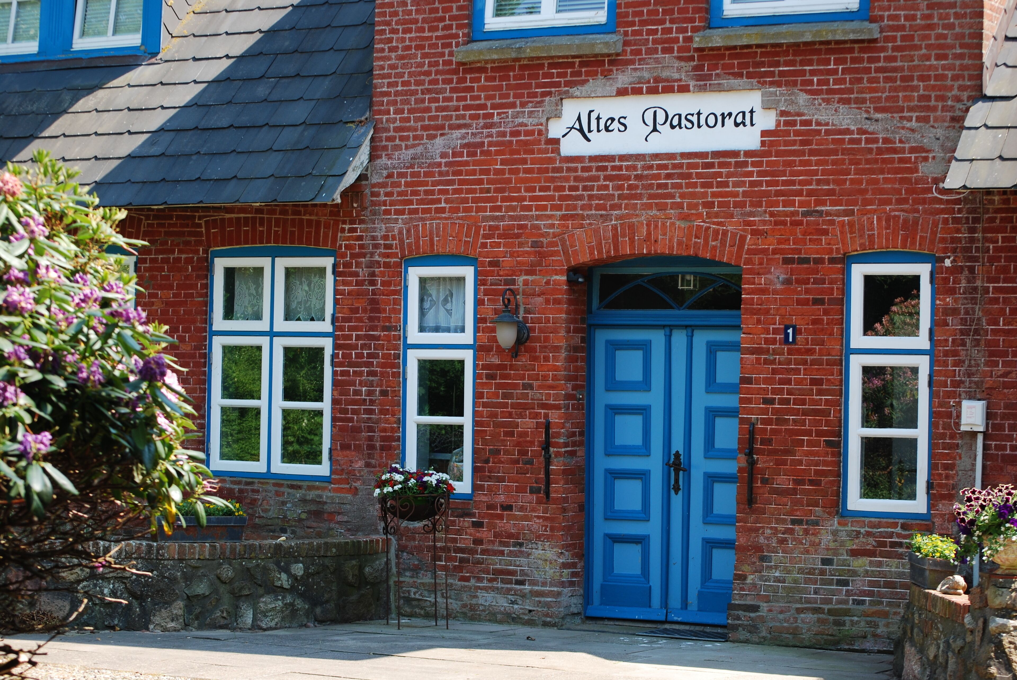 Ein rot backsteinsichtiges Gebäude mit blauer Tür und Fenstern, mit der Aufschrift "Altes Pastorat".