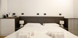 A hotel room with a bed, two bedside lamps, a small table with a lamp, a heater, and a wall-mounted control panel.