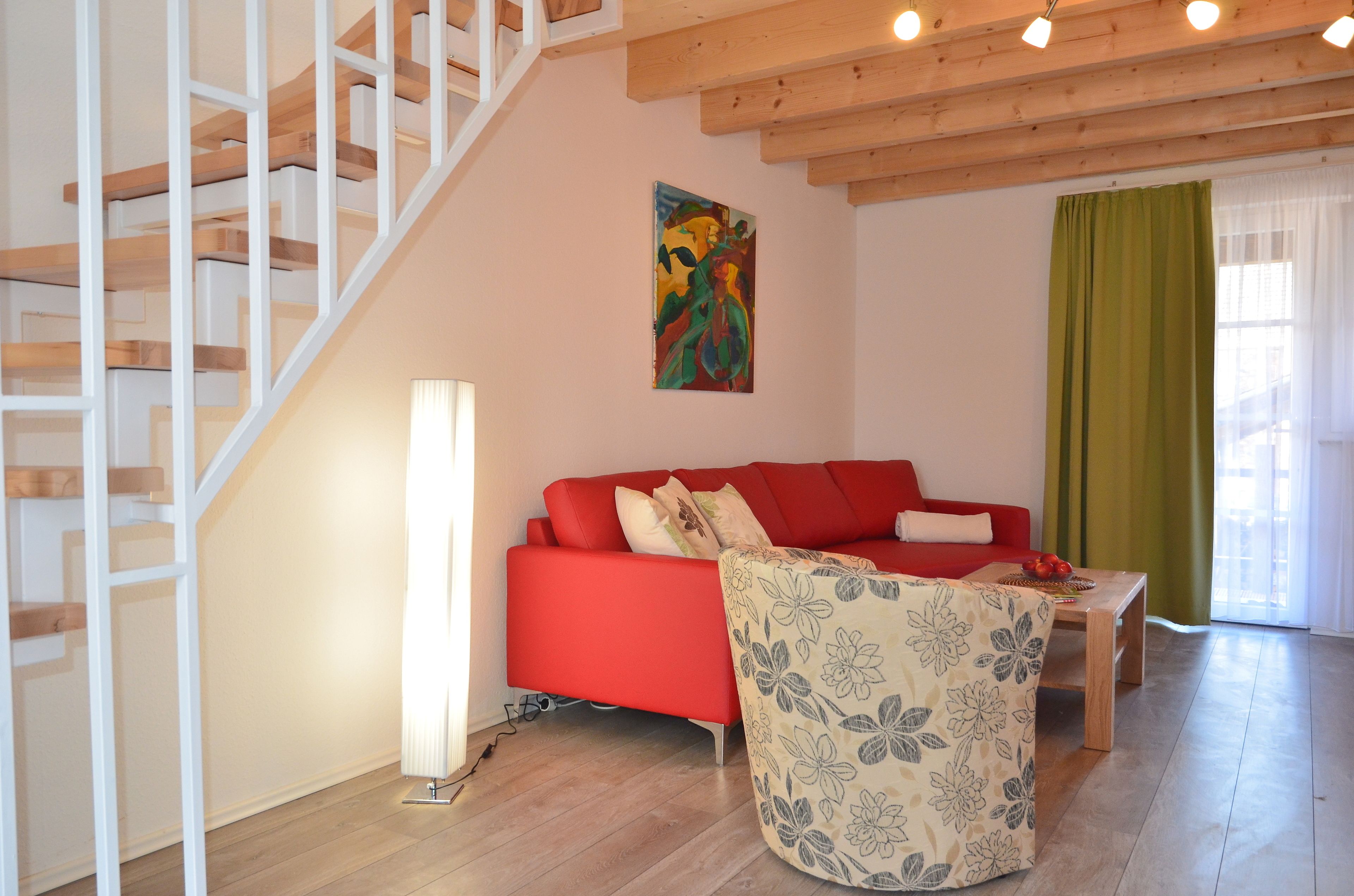 A living room with a red sofa, a wooden coffee table, a floral armchair, a staircase, and a painting on the wall.