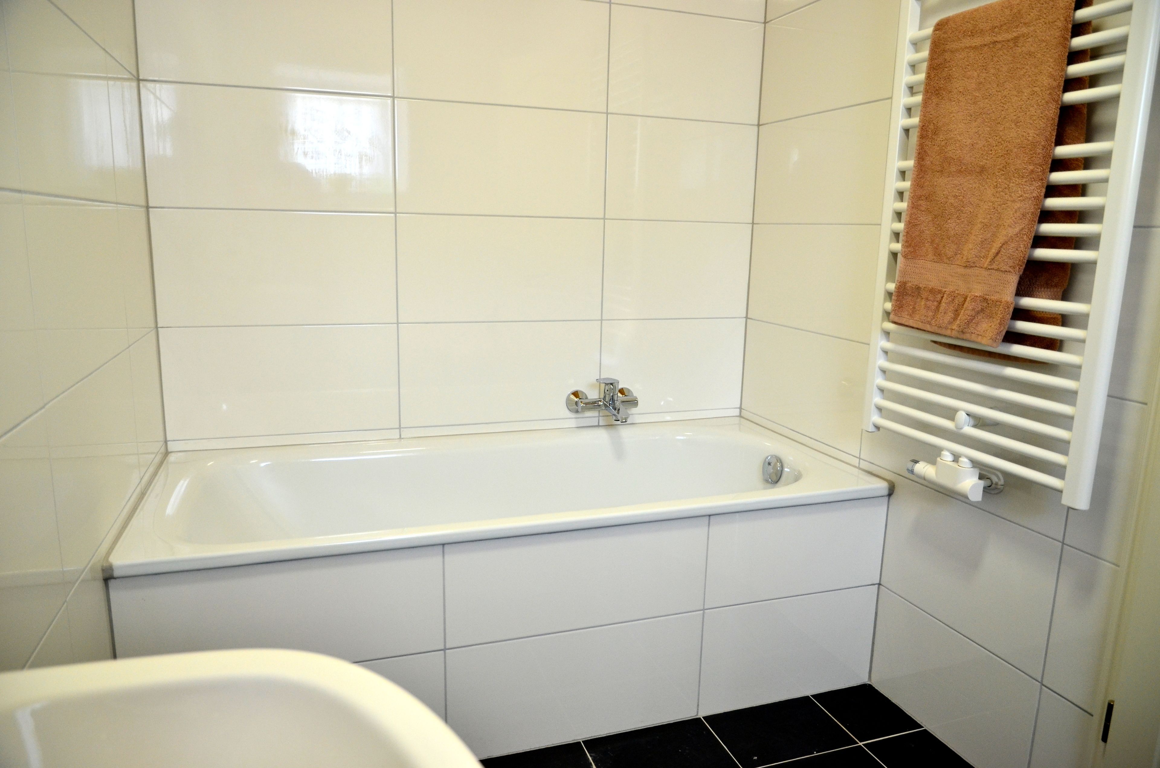Un bagno con piastrelle bianche, una vasca da bagno bianca e un portasciugamani con un asciugamano marrone.
