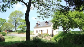 Une maison blanche avec un toit en pente, entourée d’une végétation luxuriante et d’arbres.