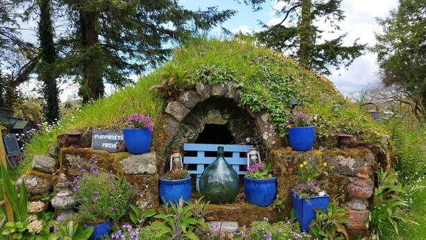 Une construction rustique en pierre, avec un toit recouvert de mousse, entourée de plantes et de fleurs en pot.