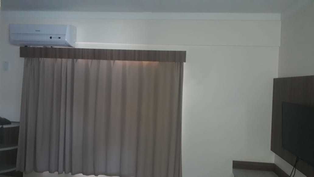 A room with a large window covered by a curtain, a flat-screen TV mounted on the wall, and an air conditioning unit above the window.