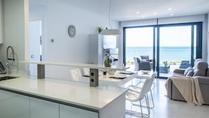 A modern kitchen with a central island, a dining area with white chairs, and a sofa with a view of the sea. - Myflats Infinity View (Elche)