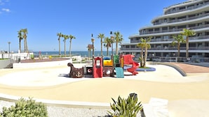 A playground with various play structures, palm trees, and a modern hotel building in the background. - Myflats Infinity View (Elche)