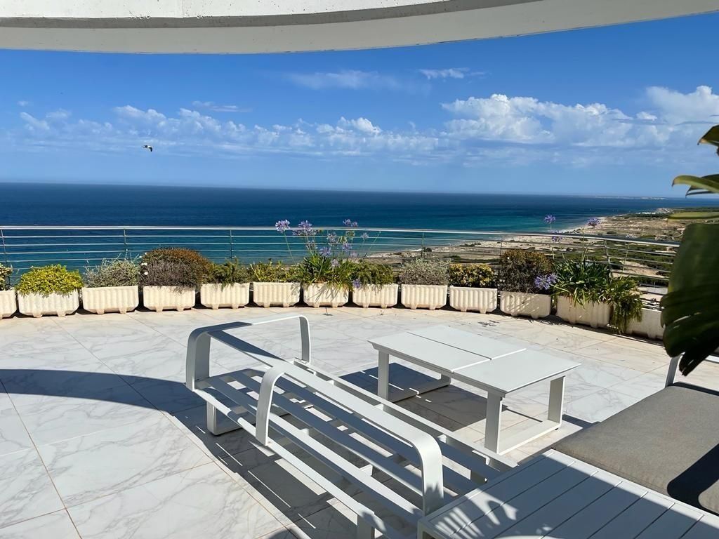 A terrace with a view of the ocean, white outdoor furniture, and potted plants.