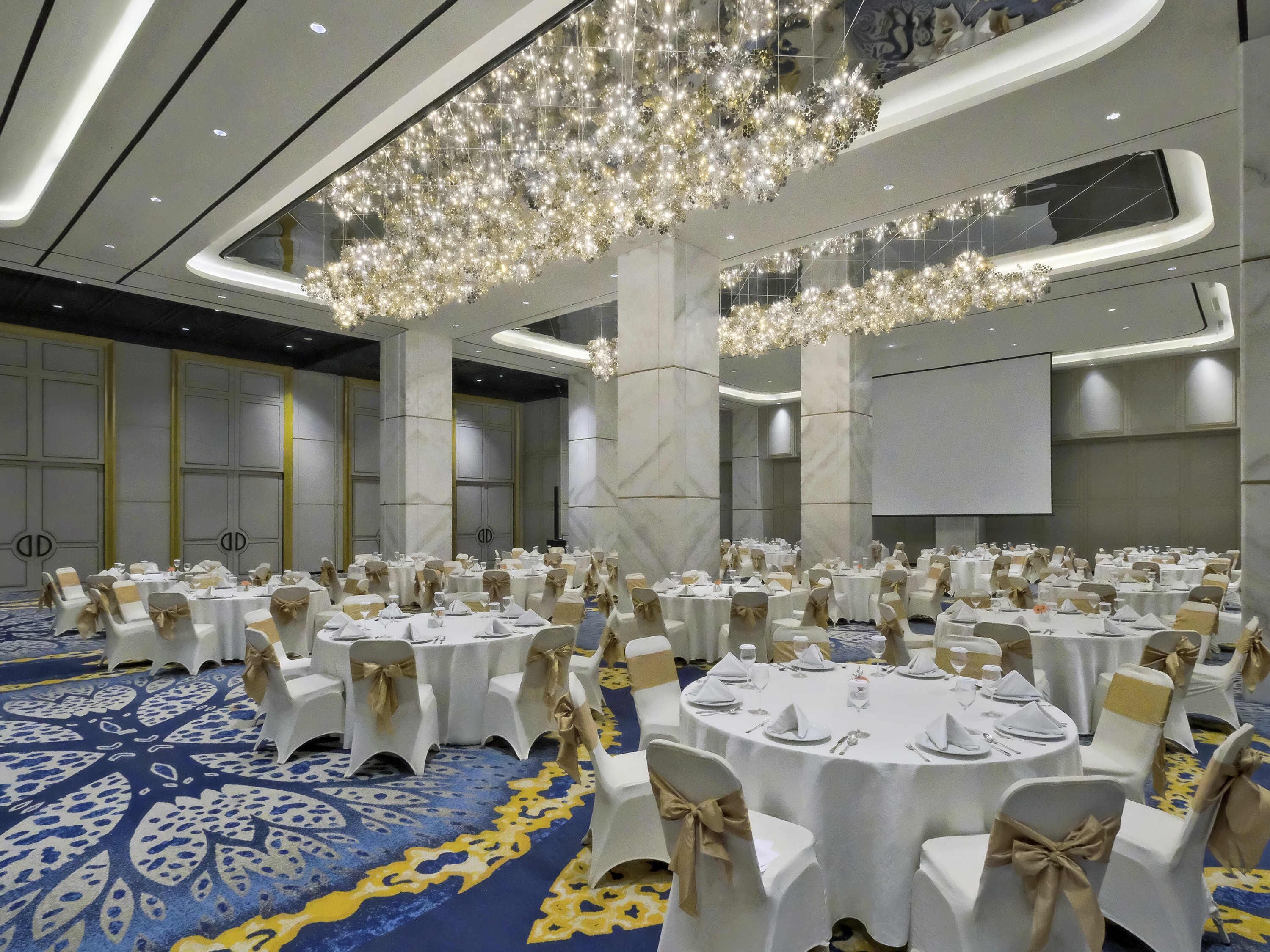 A banquet hall with round tables, white chairs with gold sashes, and a blue patterned carpet.