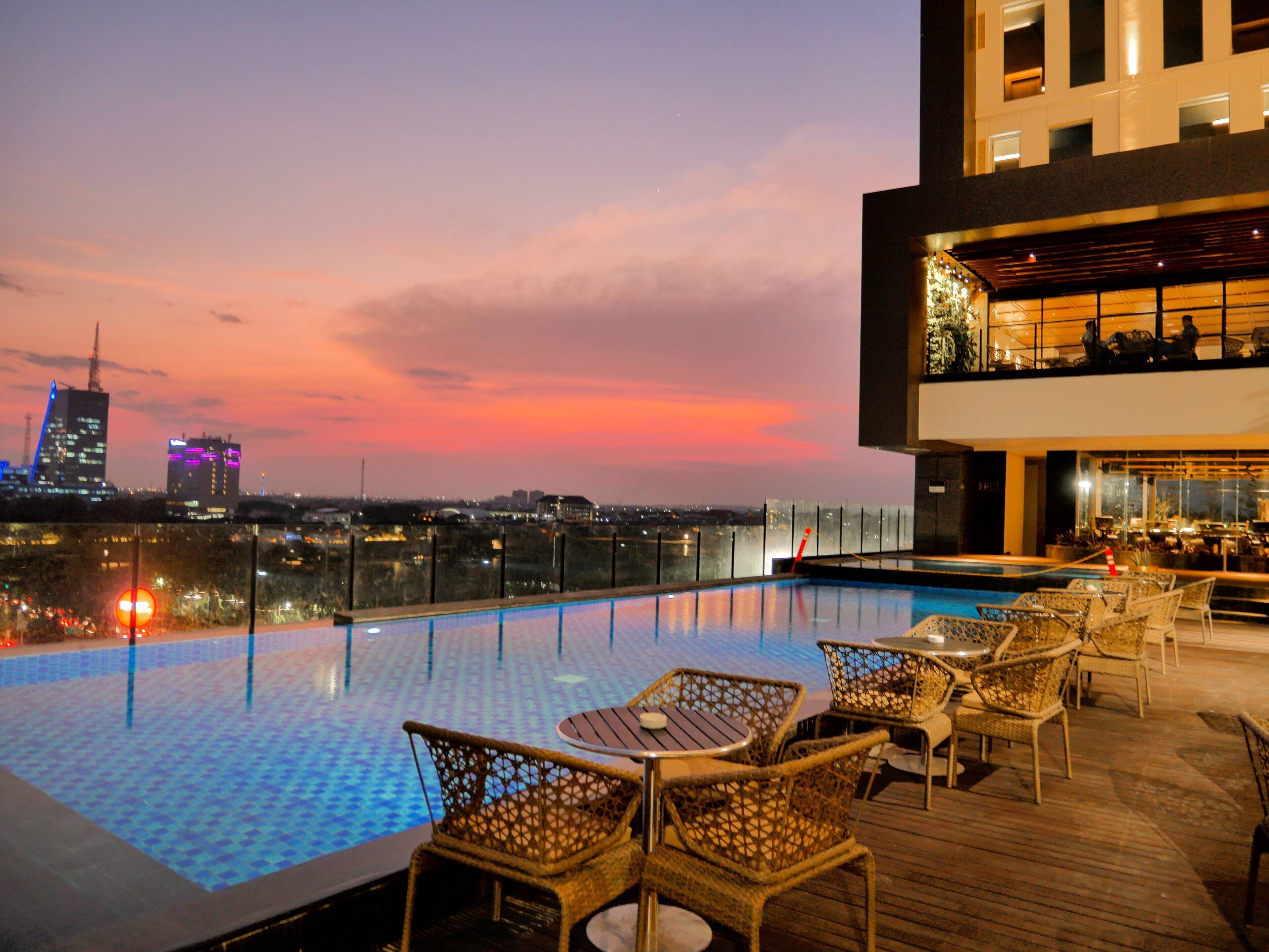 A rooftop pool with a city view at dusk, featuring a bar area with wicker chairs and a modern building in the background.