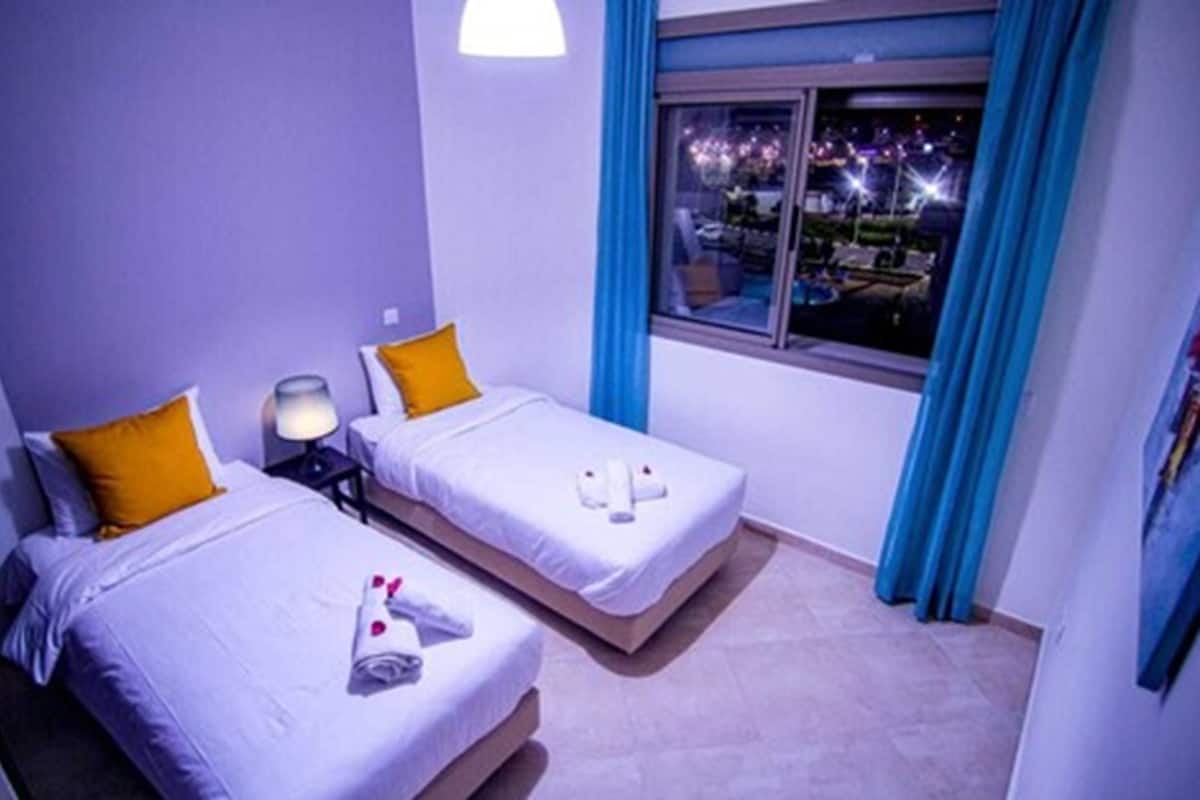A hotel room with two beds, each with a pillow and a small table with a lamp. There is a window with curtains and a view of the outside.