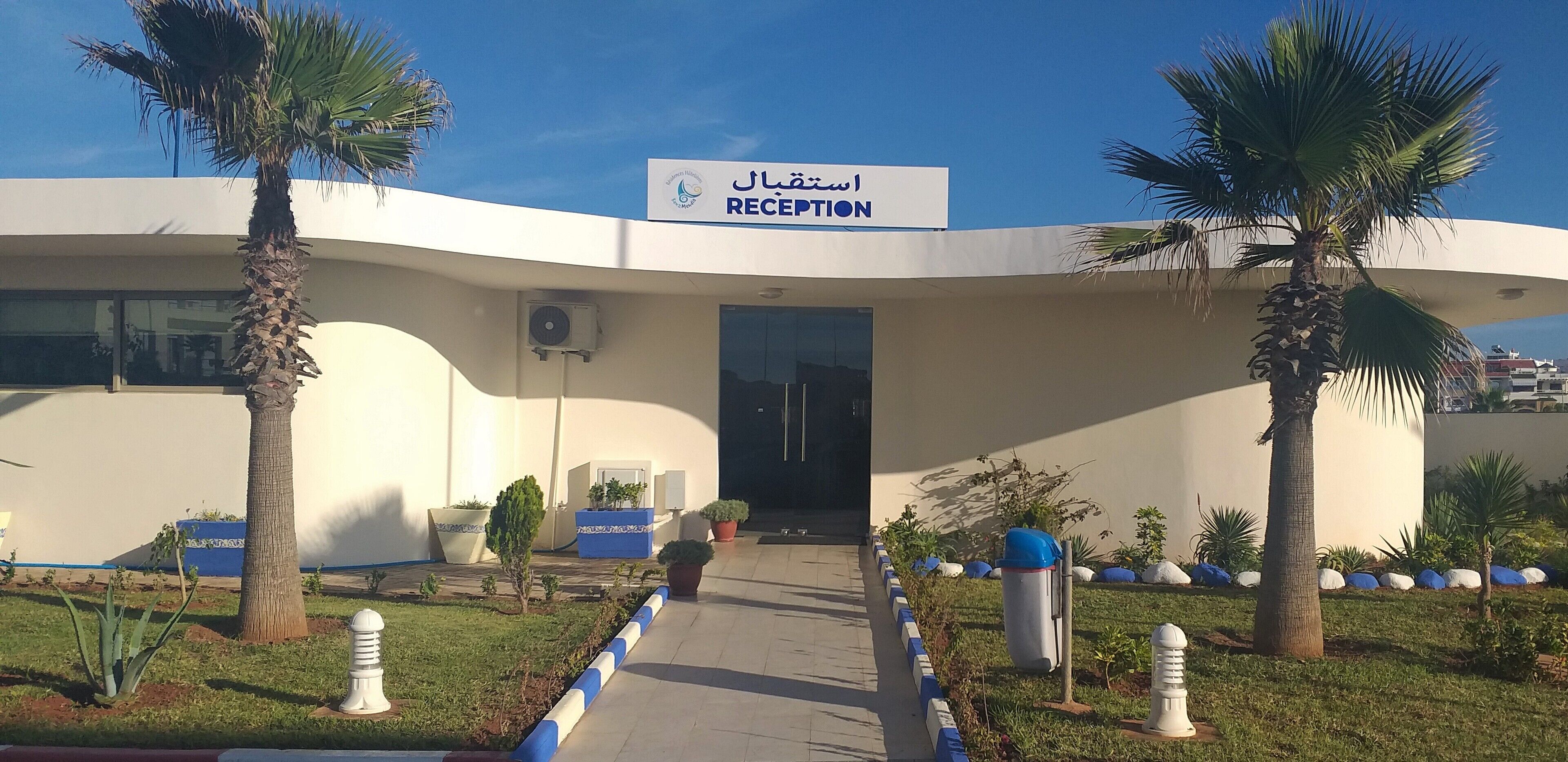 A reception area with a sign in Arabic and English, palm trees, and a pathway leading to the entrance.