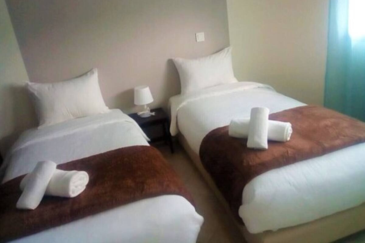 A hotel room with two beds, each with white linens and brown throw blankets. There is a nightstand with a lamp between the beds.