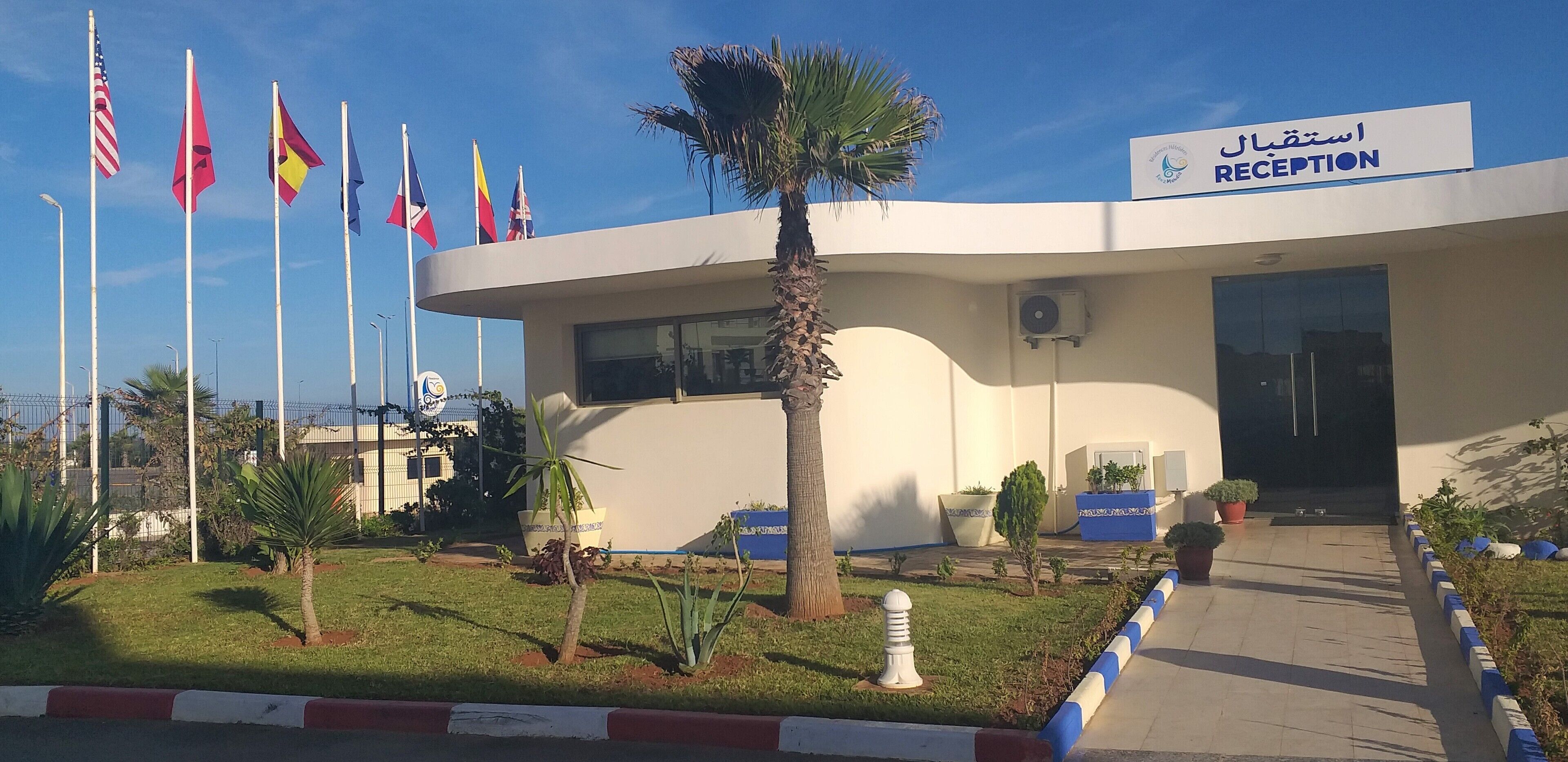 Ein Empfangsgebäude mit Schilder in Arabisch und Englisch, umgeben von einem gepflegten Garten mit verschiedenen Pflanzen und einer Palme.